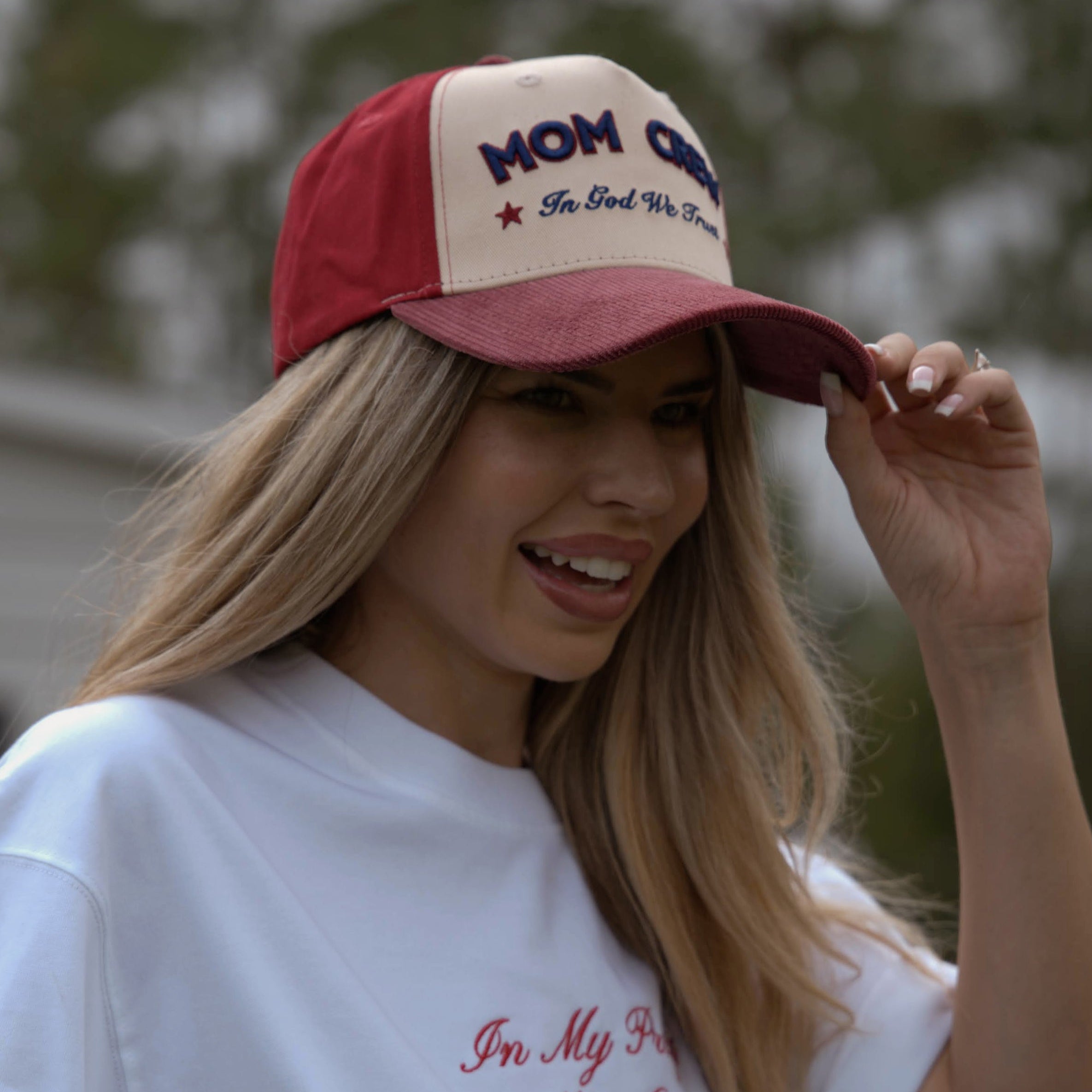 Woman wearing a red and white cap and white t-shirt with text, outdoors.